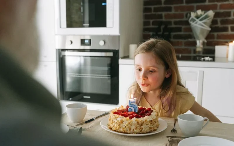A young girl blows out a birthday candle on a cake during a fifth birthday celebration indoors.