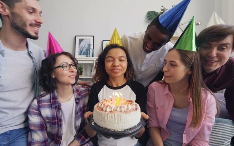 Group of friends celebrating a birthday with a cake and party hats indoors.