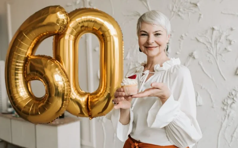 Senior woman with silver hair joyfully holding a cupcake next to 60 balloon indoors.