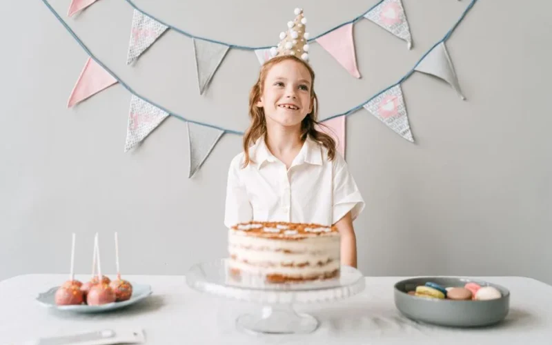A smiling girl at a birthday party with a cake, wearing a party hat indoors.