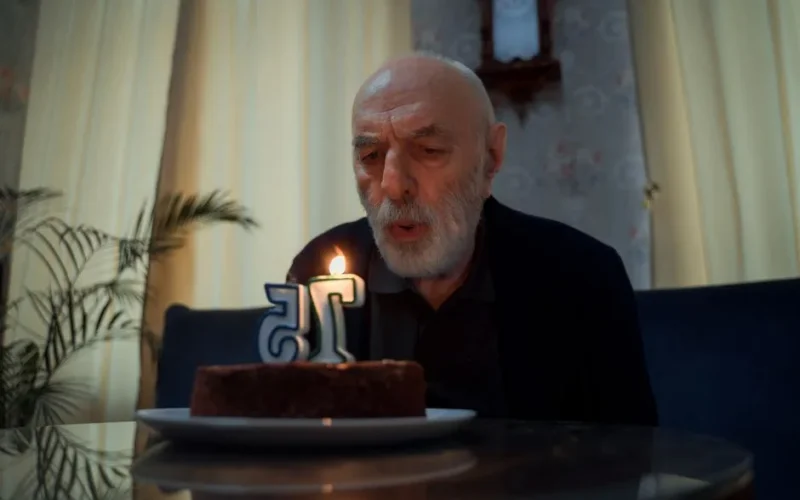 Senior man blowing candle on a 75th birthday cake indoors.