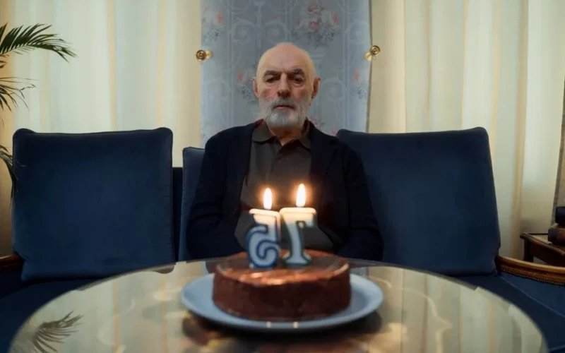 A senior man sitting in a living room with a birthday cake and candles on a glass table.