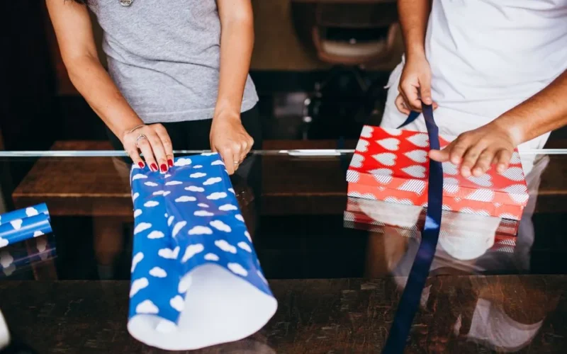 Two adults wrapping gifts with colorful paper and ribbons indoors.