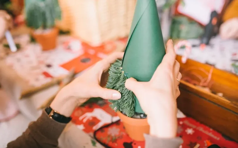 Hands crafting a DIY Christmas tree using green paper and pine leaves for holiday decor.