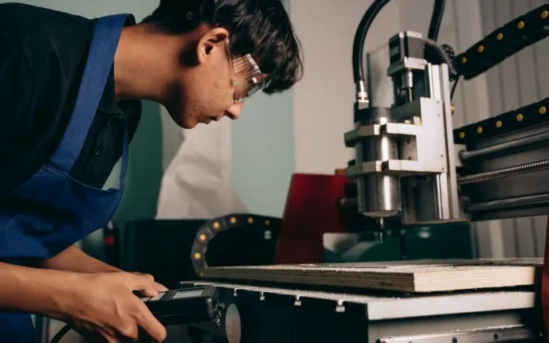 A focused young man wearing safety goggles operates a CNC machine in an industrial workshop.