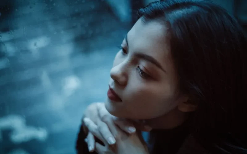 A thoughtful woman looks out a rainy window, deep in contemplation.