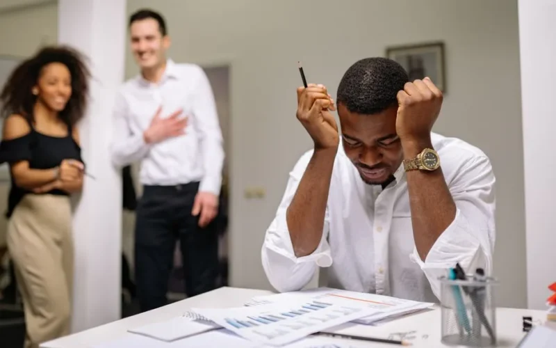 Stressed man at an office desk surrounded by colleagues, feeling overwhelmed at work.