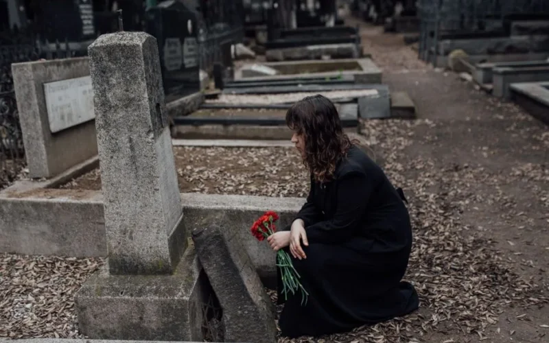 A woman in mourning holds red flowers in a quiet cemetery.