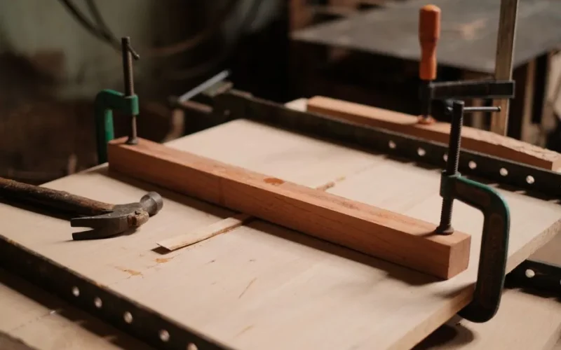 A woodworking workshop scene featuring clamps, a hammer, and wooden planks on a table.