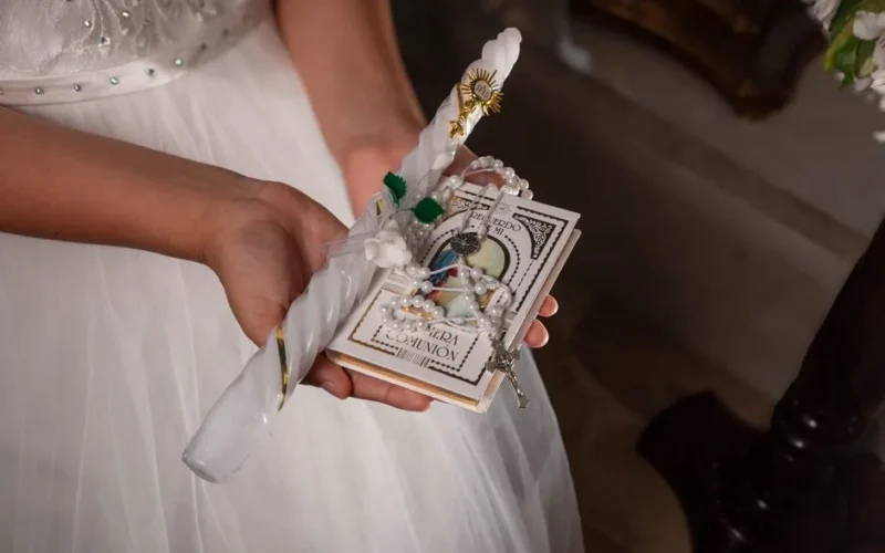 Close-up of hands holding First Communion items including candle, rosary, and book.