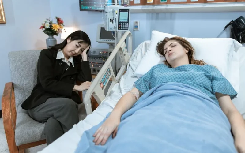 A woman sits by a hospital bed, looking worried, as a patient rests peacefully.