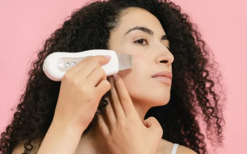Young woman with curly hair applying a cavitation peeling device, enhancing skincare routine.