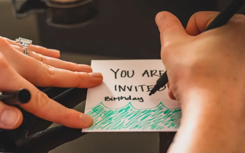 Hands writing a personalized birthday invitation card using a marker on paper.