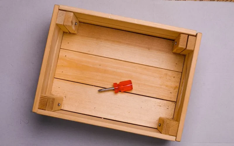 Flat lay of a red screwdriver inside an empty wooden box on a gray background.