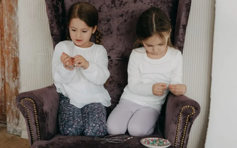 Two young girls sitting on a velvet chair crafting beaded bracelets with colorful beads indoors.
