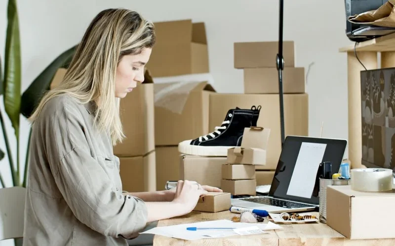 A woman packing boxes for her online store, surrounded by packaging materials and a laptop.