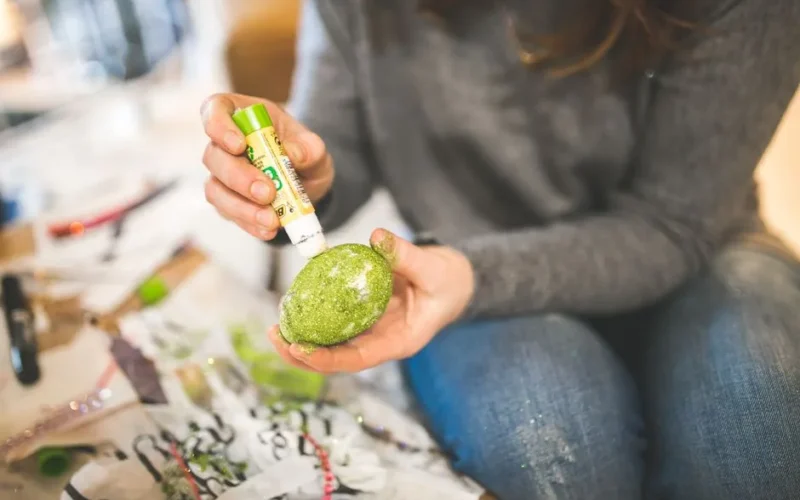 A close-up of a woman decorating an egg with green glitter at home.