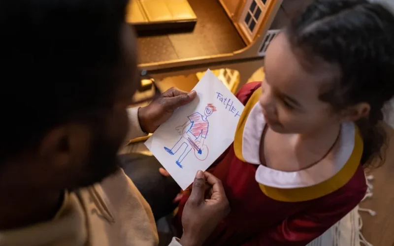 A father and daughter share a touching moment over a child's drawing labeled 'Father'.