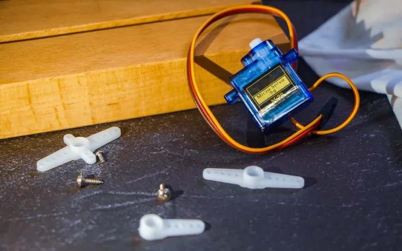 Close-up of a micro servo motor with plastic arms and screws on a workbench, ideal for robotics projects.
