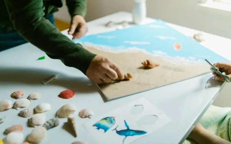 Hands working on a seashell artwork on a table with scattered seashells and other art materials.