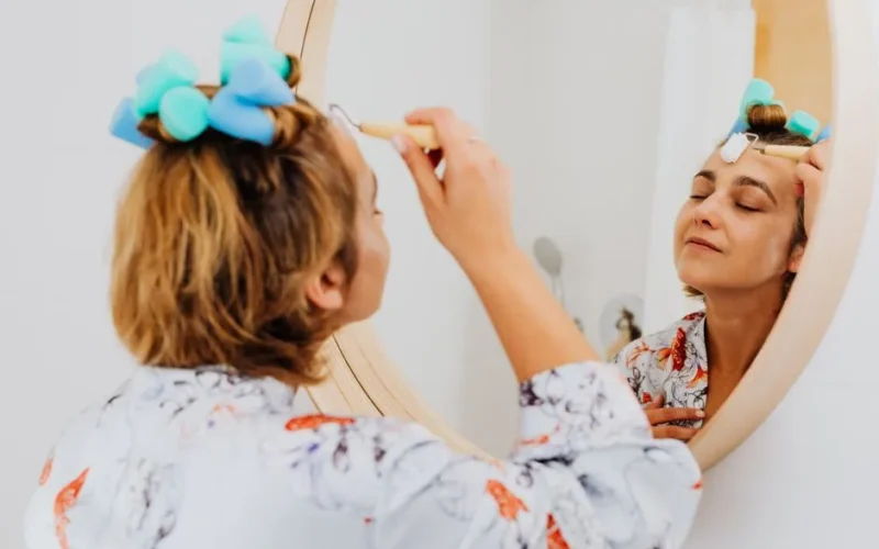 Woman with closed eyes applying skincare product in front of mirror, enjoying self-care time.