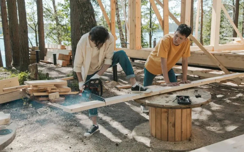 Two adult men working on wood construction project outdoors, using a chainsaw.