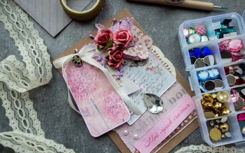 Top view of handicraft greeting card decorated with artificial flowers and fancy stones placed on table near box of colorful beads and buttons and tape lace