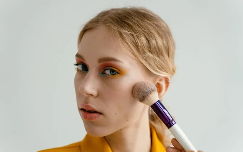 A woman applying blush with a makeup brush, showcasing a colorful eyeshadow look.