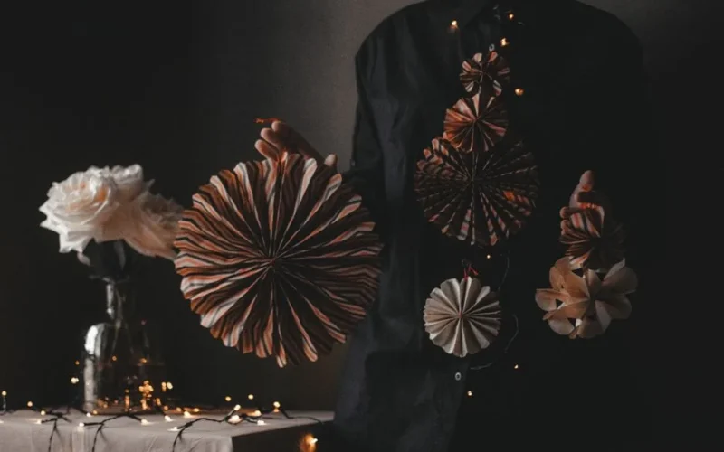 Unrecognizable female with various creative decorations made of paper standing at table with garland and flowers on dark background in room