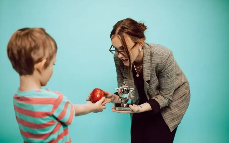A child offers an apple to a teacher holding a "Best Teacher" trophy in a classroom scene.
