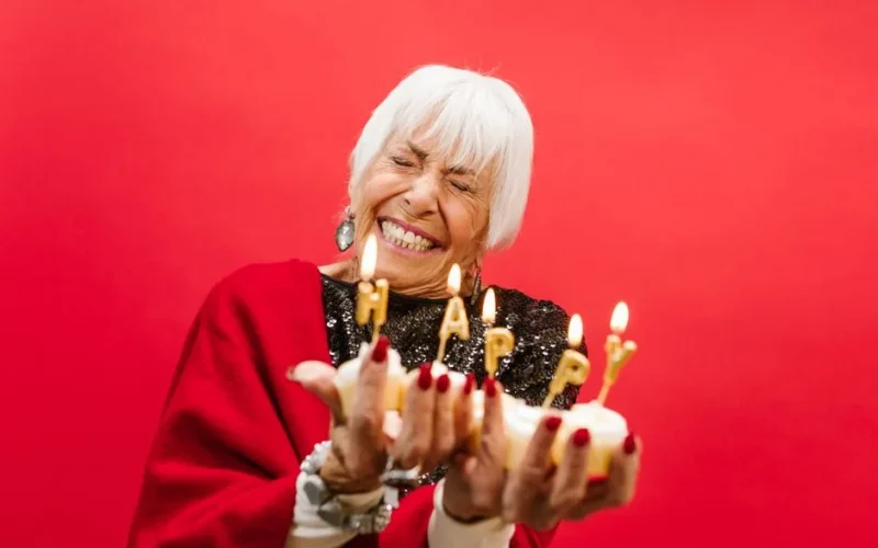 Cheerful elderly woman holding a birthday cake with lit candles spelling 'HAPPY' against a red background.