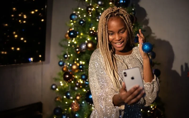A joyful woman taking a selfie in front of a decorated Christmas tree with her smartphone.