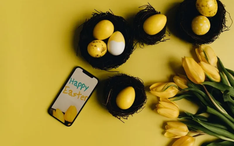 Yellow Easter eggs and tulips beside smartphone with 'Happy Easter' on yellow background.