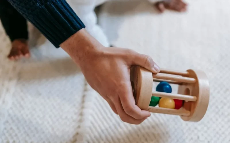 From above of crop anonymous male parent carrying colorful wooden rattle with small balls while spending time with little babyb