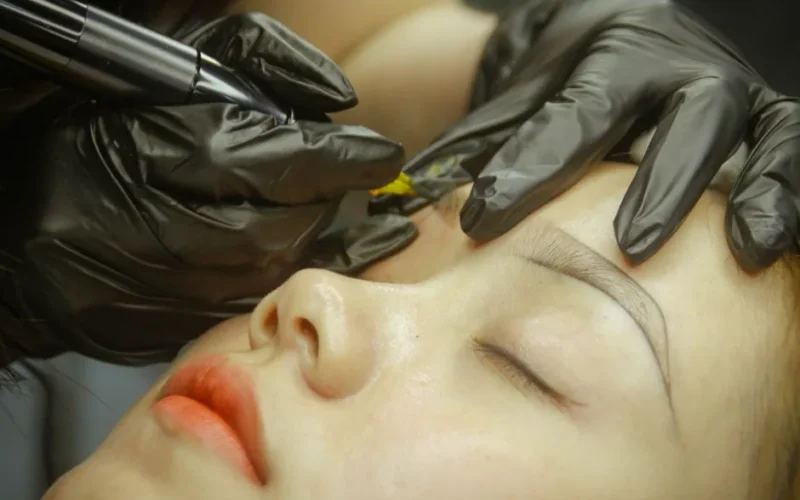 A detailed view of eyebrow microblading procedure with gloved hands and closed eyes.