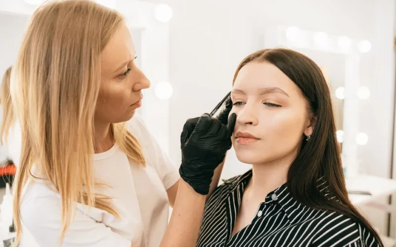 A skilled makeup artist applies eyebrow makeup on a client in a light-filled studio.