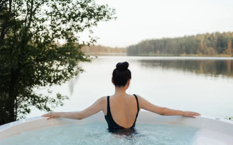 A woman relaxes in a jacuzzi outdoors, overlooking a serene lake and forest landscape.