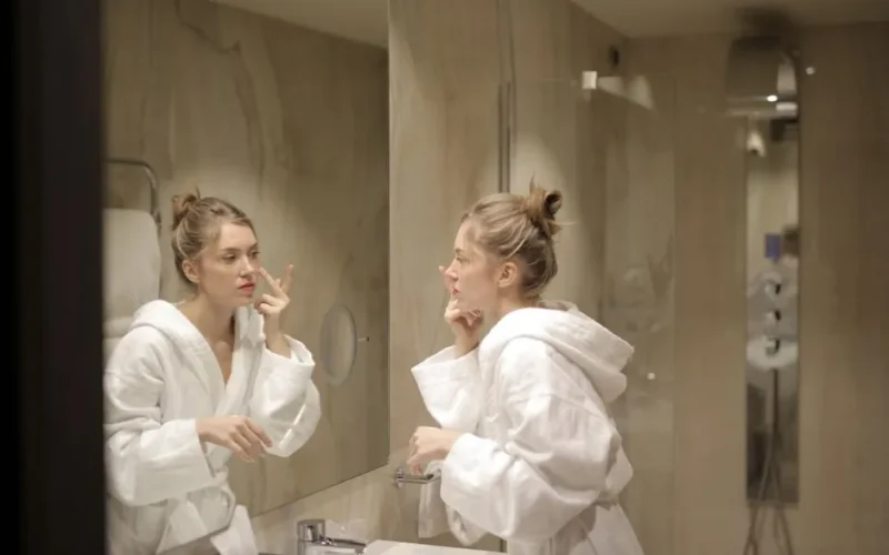A woman in a white bathrobe applies moisturizer while looking at her reflection in the bathroom mirror.