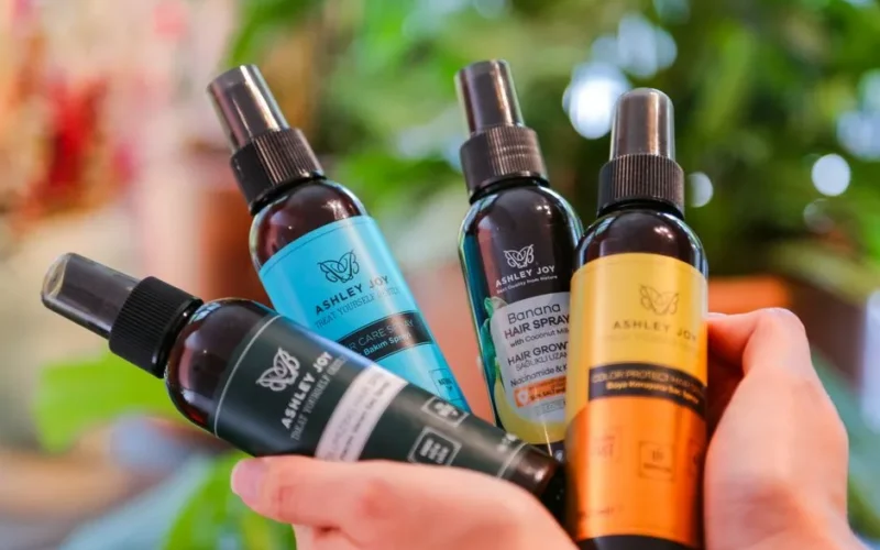 Close-up of hands holding various Ashley Joy hair care bottles with a blurred natural background.