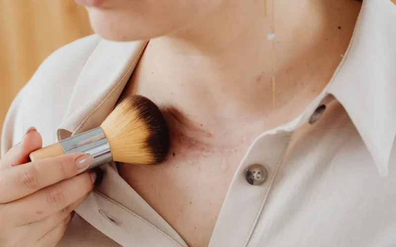 A woman uses a makeup brush to enhance her skin, blending scars for a natural look.