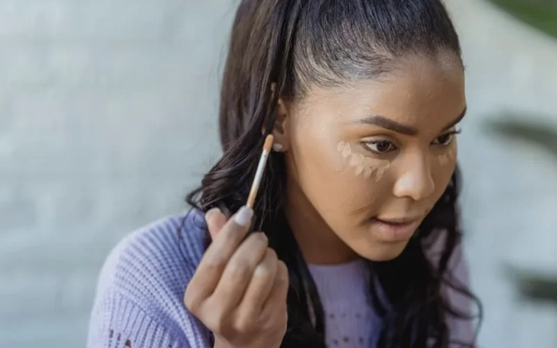 Crop African American woman with long dark hair applying concealer under eyes and looking away in light room