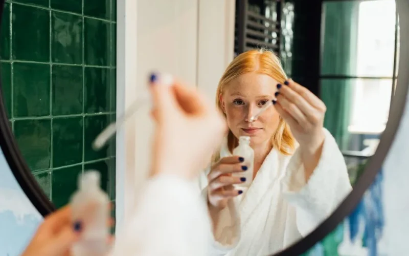 Adult woman applying skincare serum while wearing a bathrobe in front of a bathroom mirror.