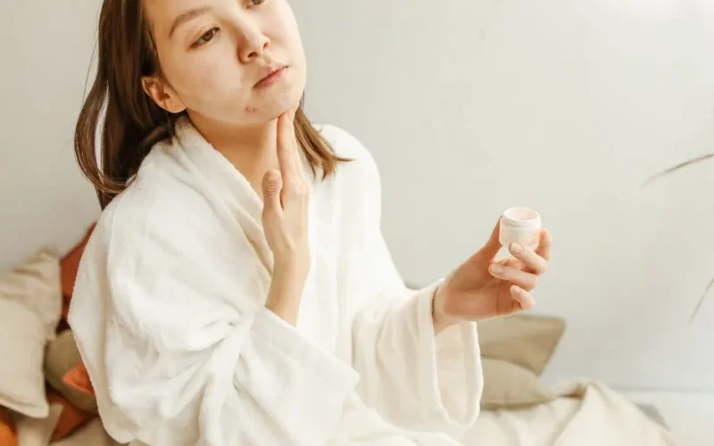 An adult woman in a bathrobe applies facial cream during her skincare routine indoors.