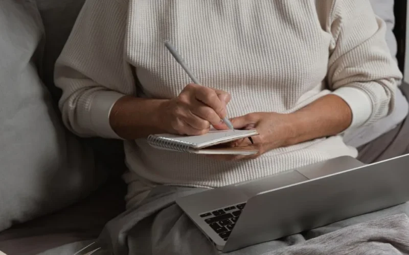 A person writing in a notebook with a pen while sitting by a laptop, creating a cozy work atmosphere.