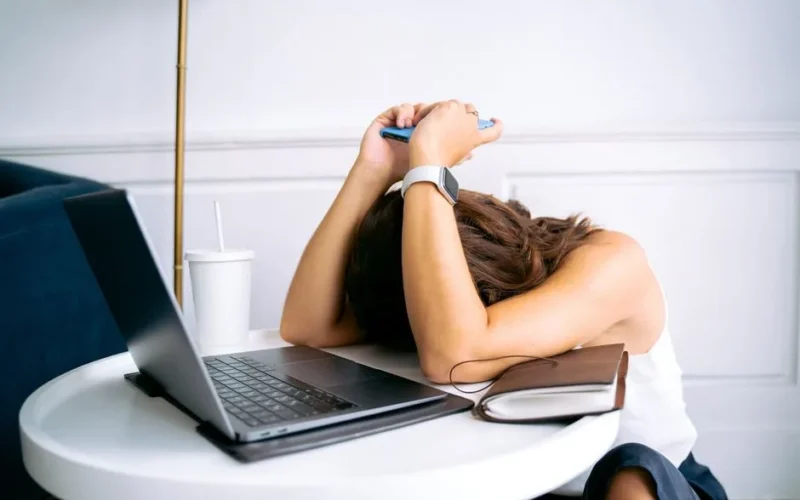 Young woman showing stress with laptop and phone at desk, embodying digital exhaustion.