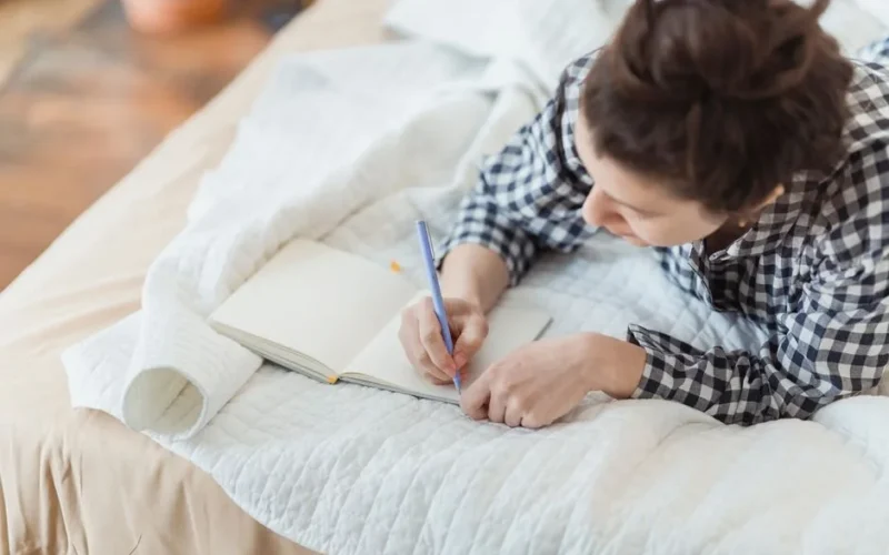 A woman writes in a journal while relaxing on a cozy bed with soft lighting and peaceful surroundings.