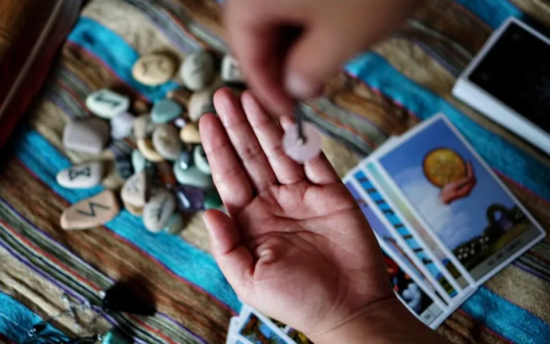 Hand holds a pendulum over runes and tarot cards, exploring divination.