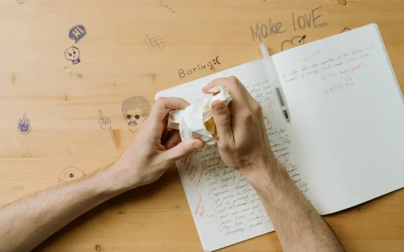 Hands crumpling paper over a notebook showing creative frustration at a writing desk.