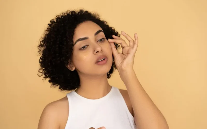 A woman with curly hair applies under eye masks against a neutral background.