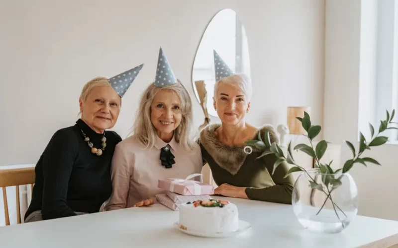 Three elderly women wearing party hats celebrating a birthday with cake and gifts.